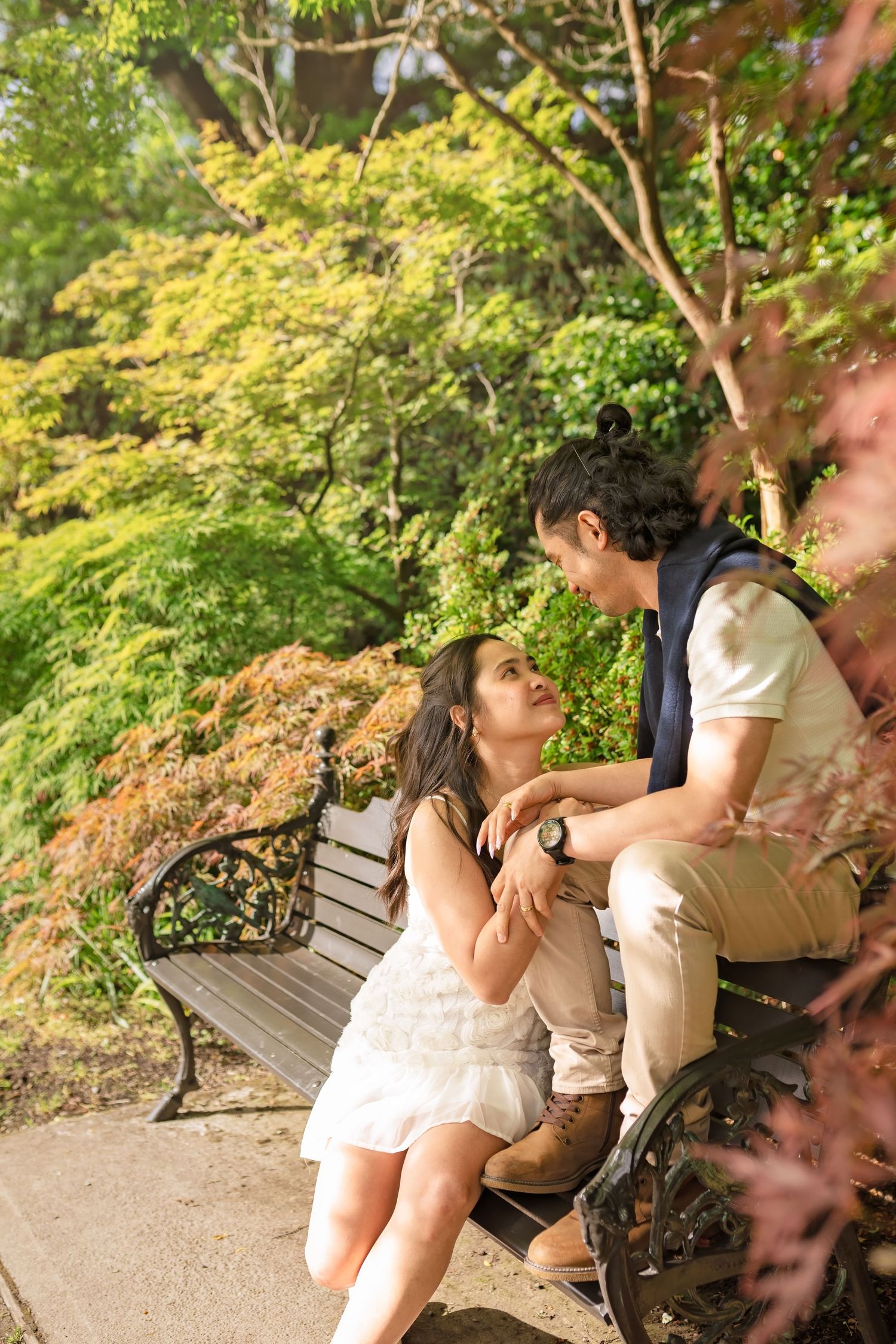 Couple sitting on a park bench surrounded by lush green and red foliage, sharing an intimate moment outdoors.