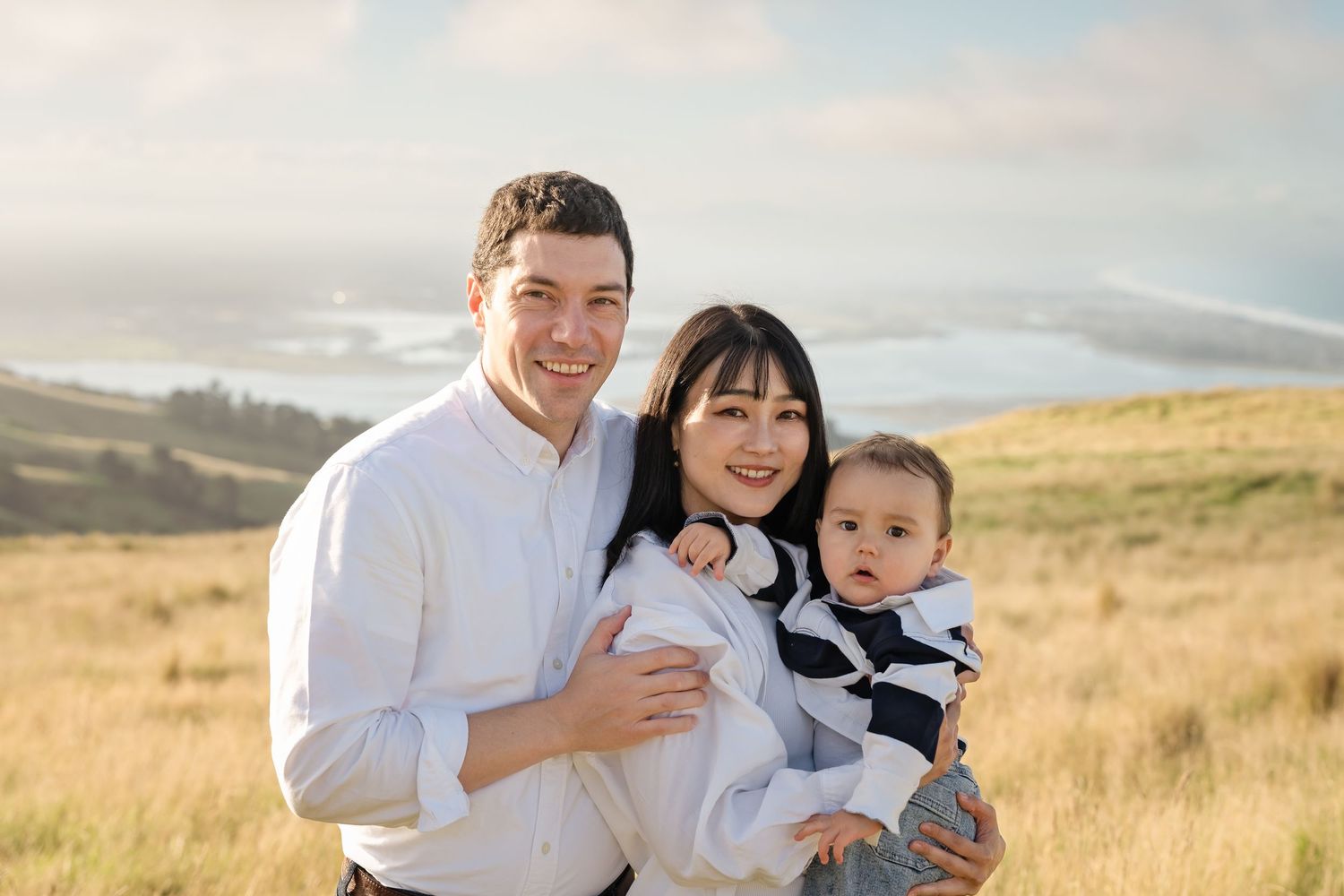 Happy family of three posing outdoors in golden field with scenic coastal hills in background.
