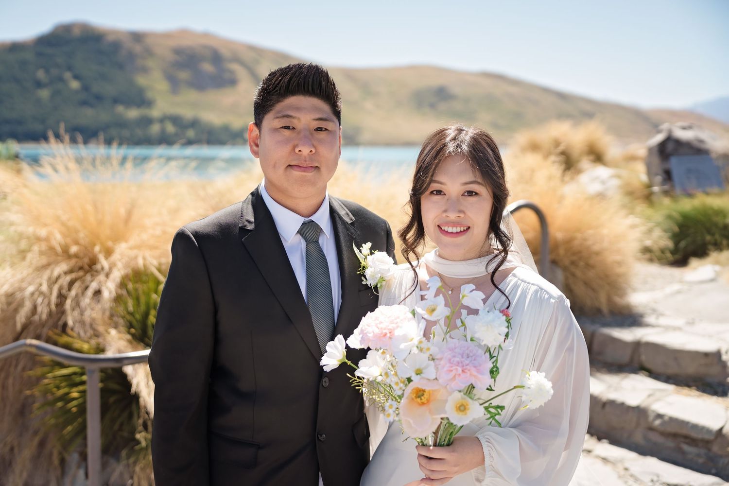Smiling bride holding a pink and white bouquet poses with groom beside a scenic lakeside backdrop.