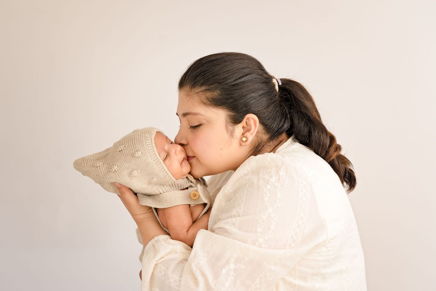 A mother tenderly kisses her newborn baby wrapped in a beige blanket against a soft white background.