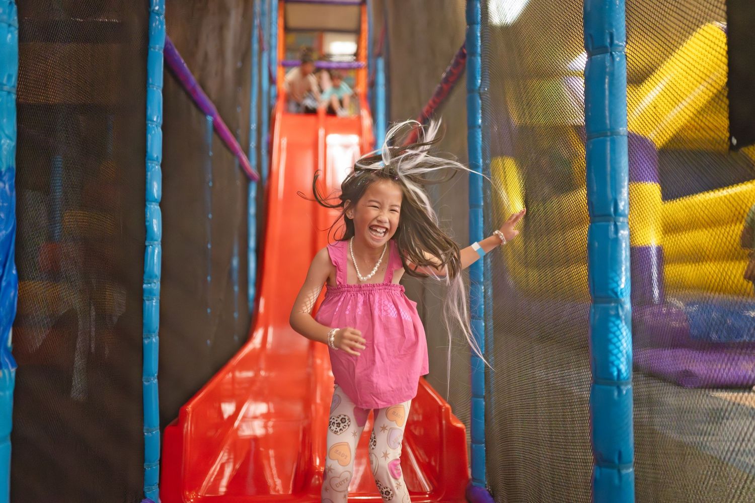 A joyful young girl in a pink dress laughs while running through a colorful indoor playground with slides.