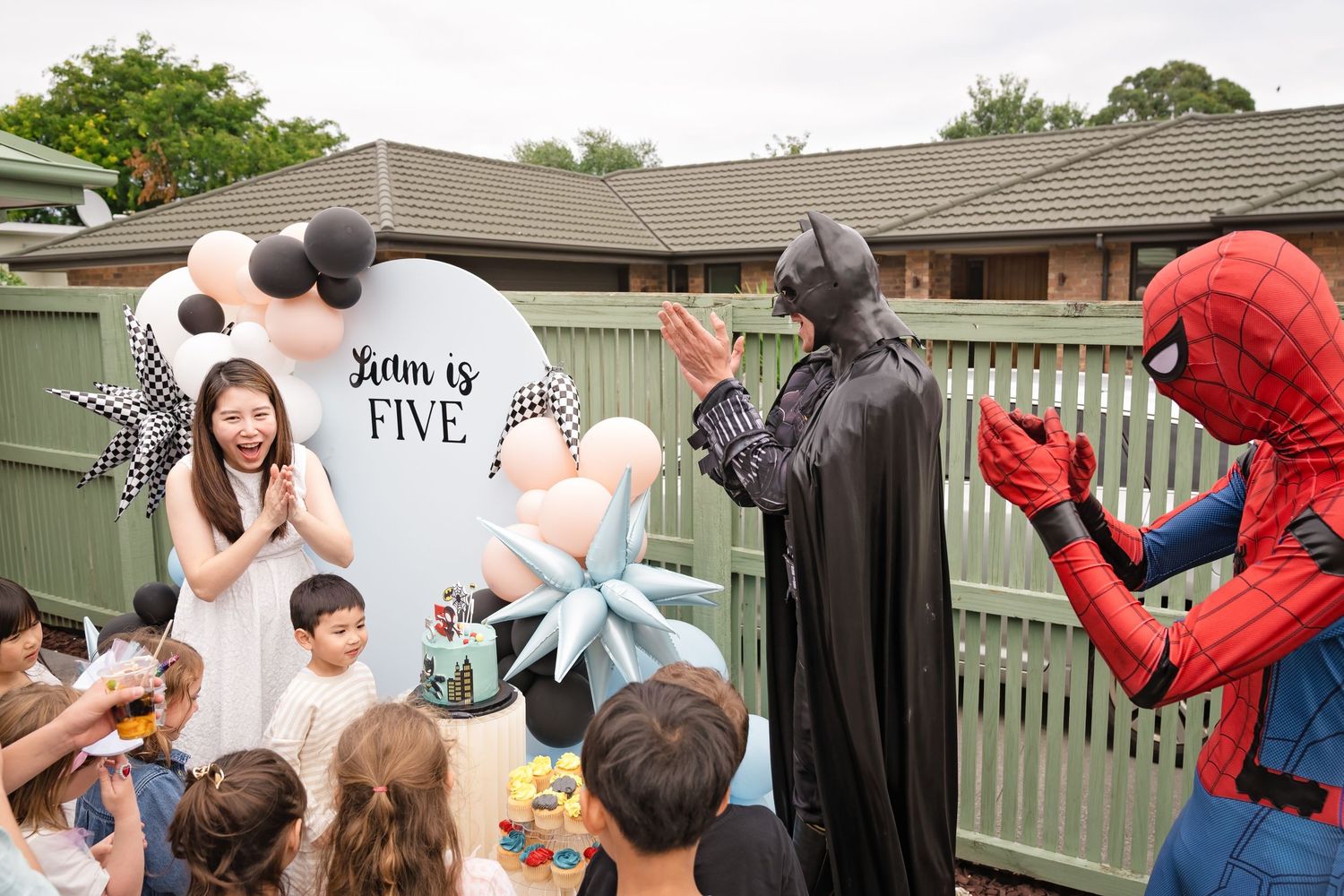Batman and Spider-Man high-five at a child's fifth birthday party outdoors with excited kids celebrating.