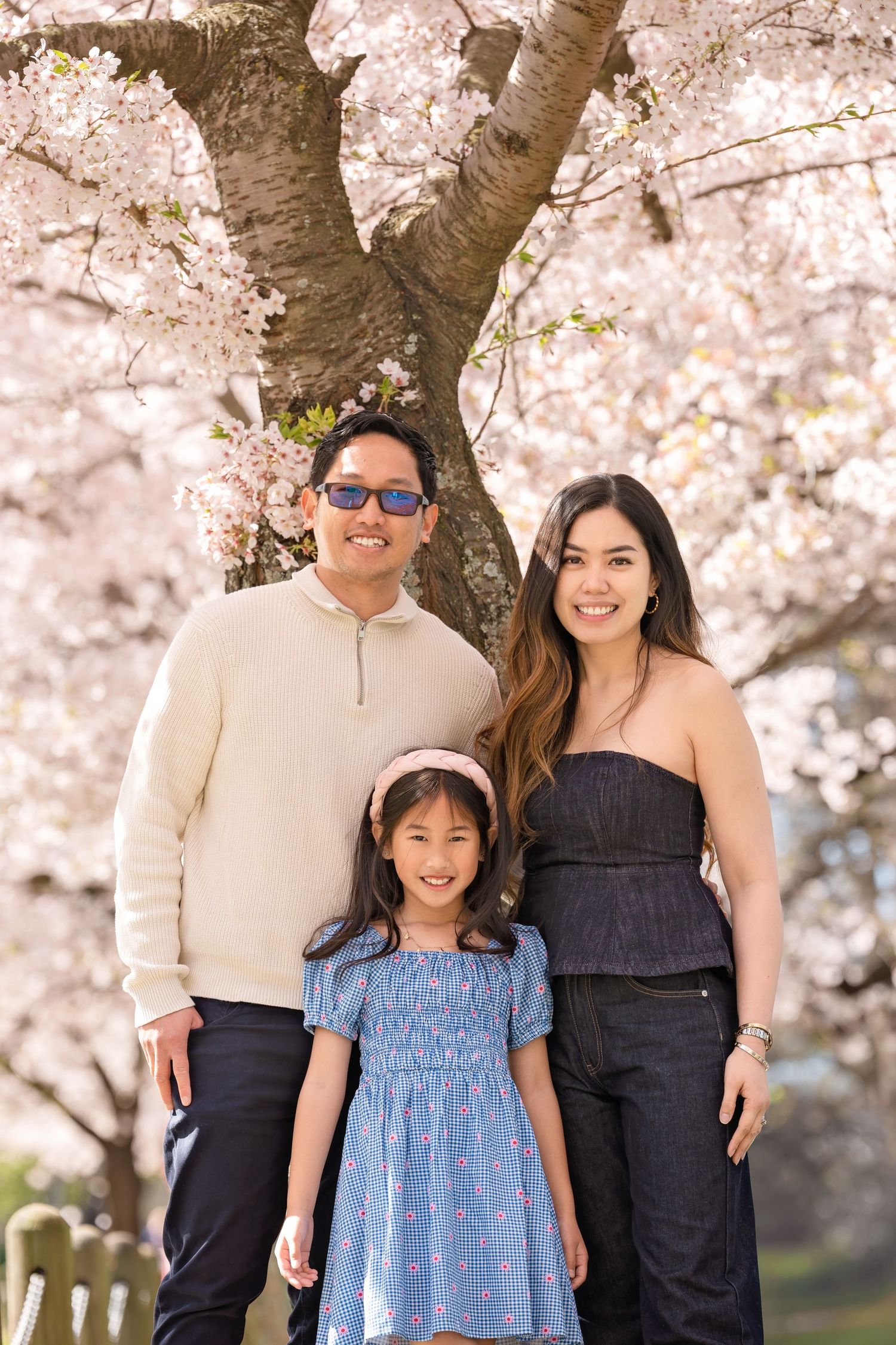 Family of three smiling under blooming cherry blossom tree in spring, parents with young daughter in blue dress.