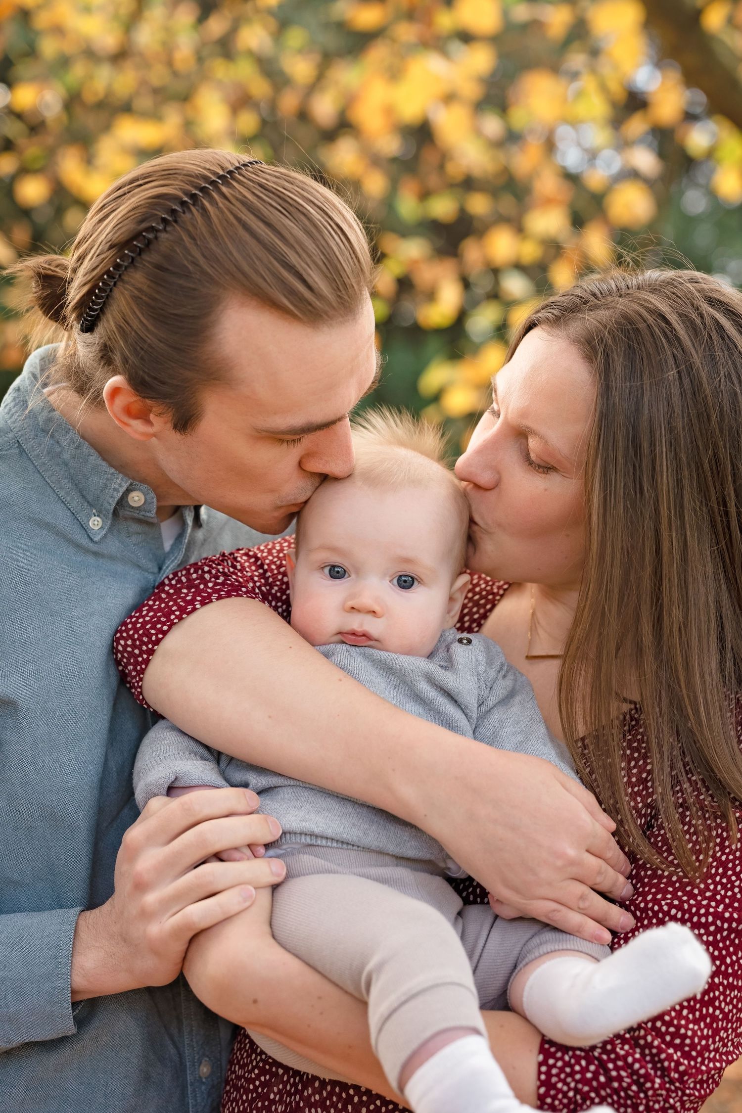 Parents kissing their baby on the cheeks outdoors with autumn yellow foliage in the background.