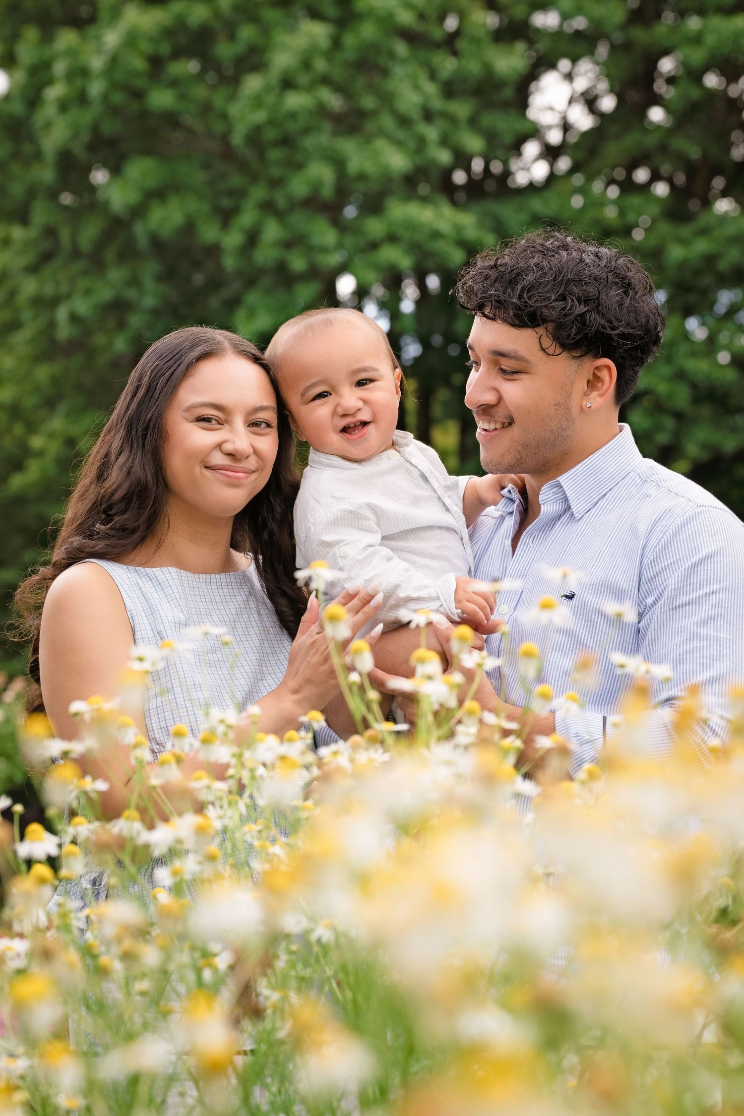 Young family with baby sitting in wildflower meadow, parents smiling at laughing infant outdoors.