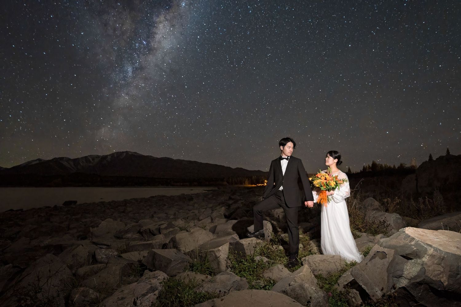 A couple in wedding attire stands on rocky terrain under a stunning starry night sky with the Milky Way visible above.