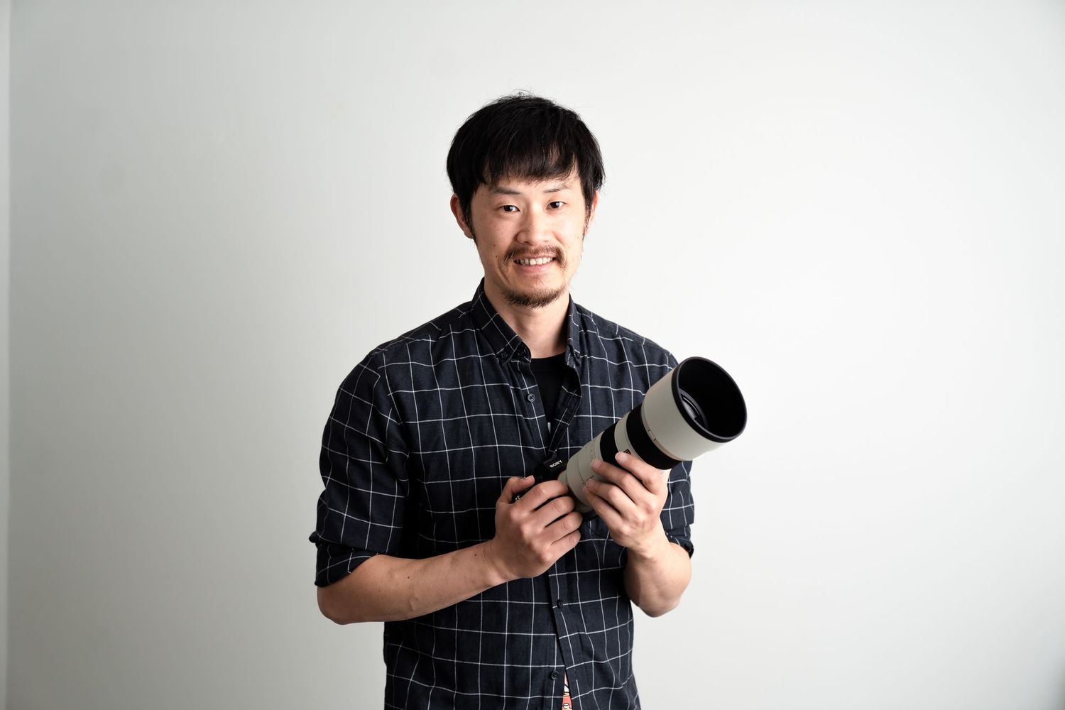 Photographer in checkered shirt holds professional camera against white background.
