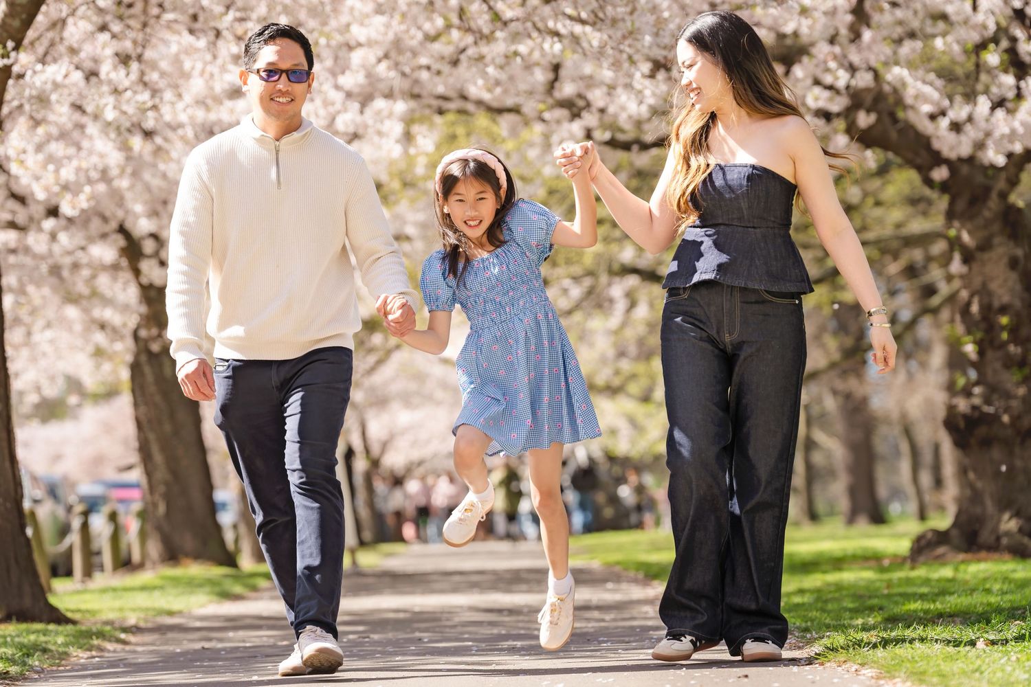 Family of three walking among cherry blossom trees, girl swinging between parents on a sunny spring day.