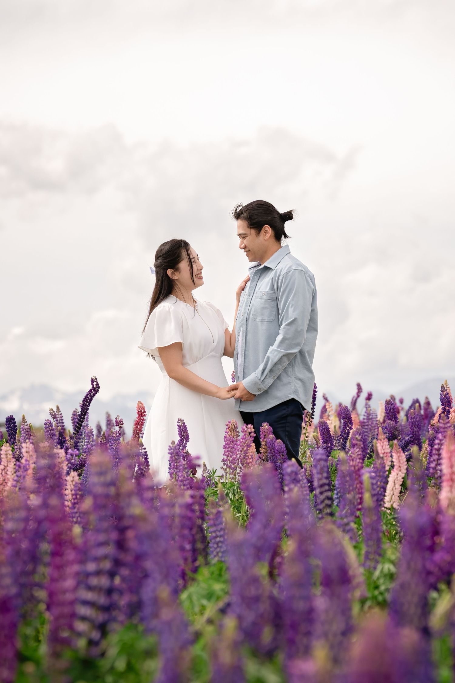 Couple holding hands and gazing at each other in a vibrant purple lupine flower field under a cloudy sky.