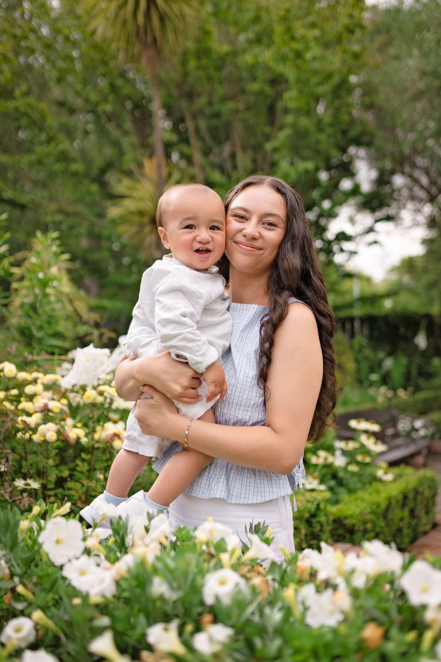 Young woman with long dark hair smiles holding a happy baby boy among white and yellow flowers in a lush garden.
