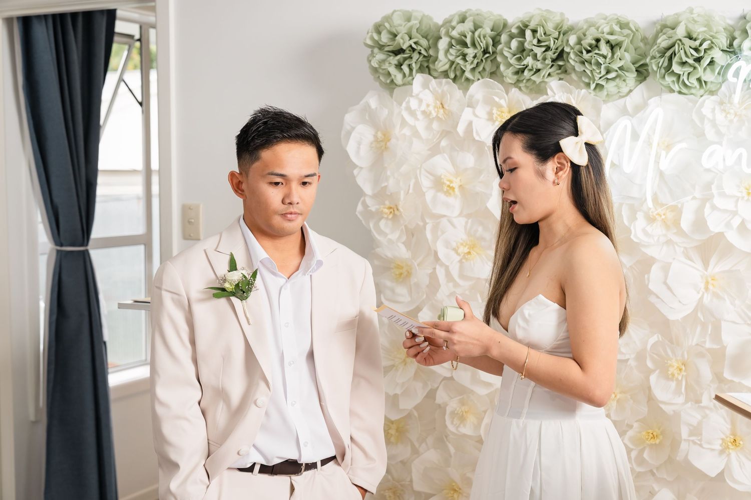 Groom in beige suit with boutonniere exchanges rings with bride in white strapless dress at wedding ceremony.