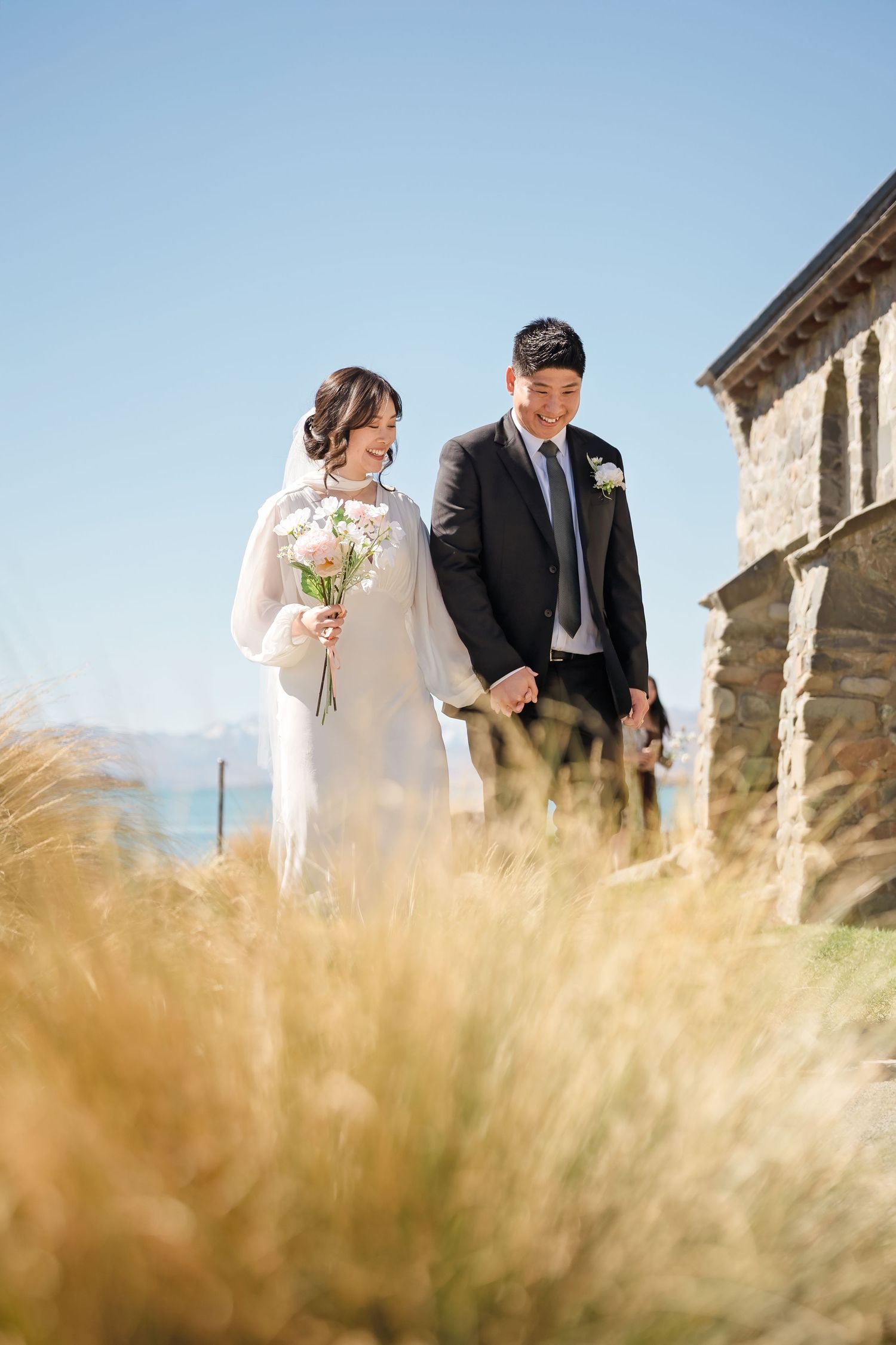 Bride holding white flowers and groom walk arm in arm past a stone chapel through golden grass.