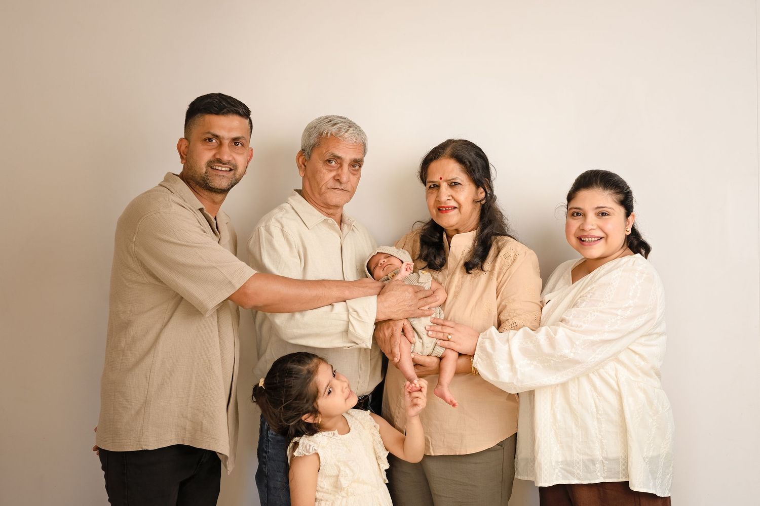 Multi-generational South Asian family posing together, holding a newborn baby, smiling warmly against a neutral background.