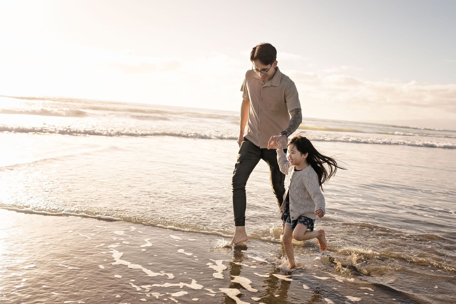A father and young daughter play together in the shallow ocean waves on a sunny beach.