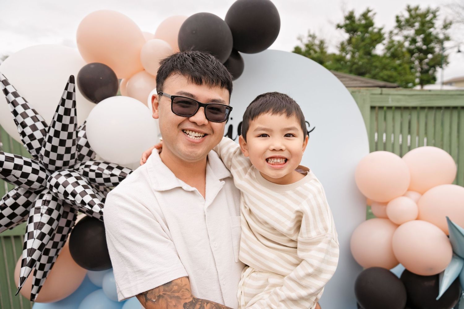 A man in sunglasses holds a smiling young boy at a Mickey Mouse balloon party backdrop.