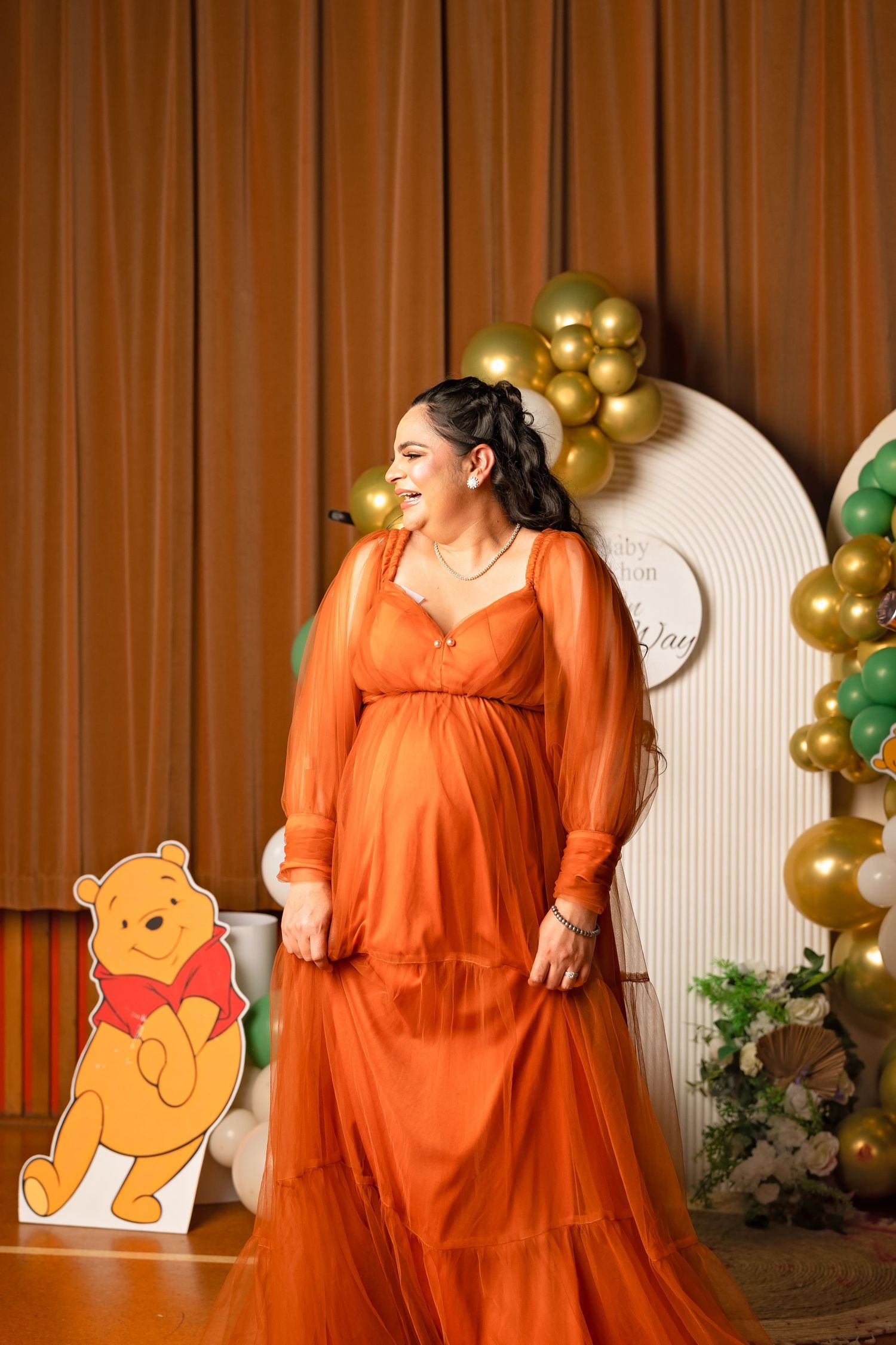 Pregnant woman in orange tulle gown at Winnie the Pooh baby shower with gold and green balloon arch.