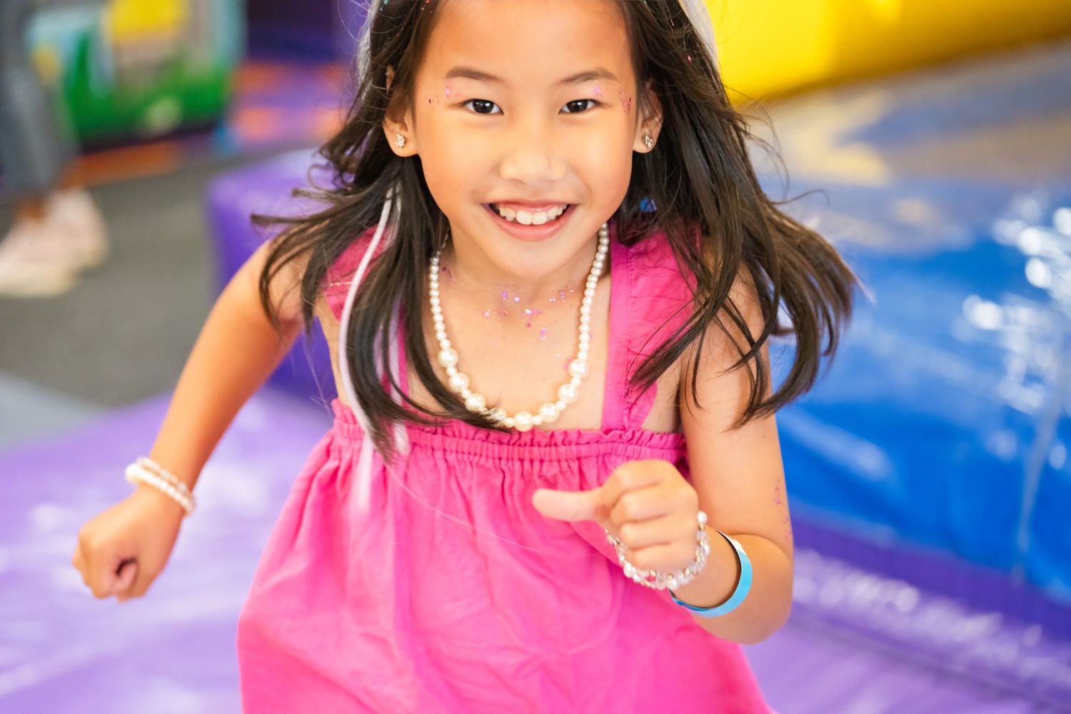 A smiling young girl in a pink dress and pearl necklace plays on colorful inflatables at an indoor bounce house.