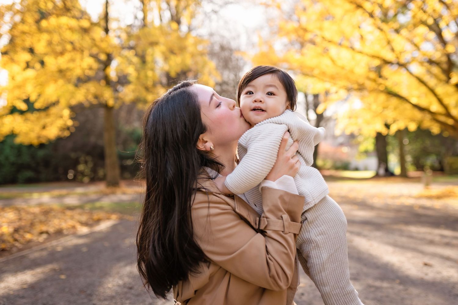 A parent and baby share a tender moment outdoors on an autumn day with golden foliage in the background.