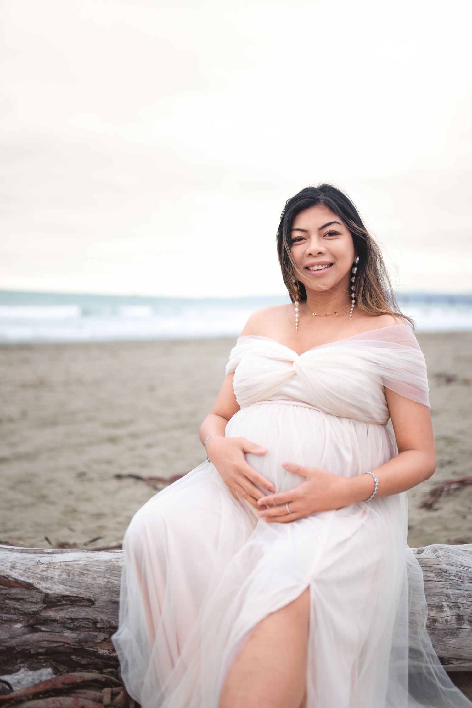 A woman in a flowing white dress poses on driftwood at the beach during a maternity photoshoot.