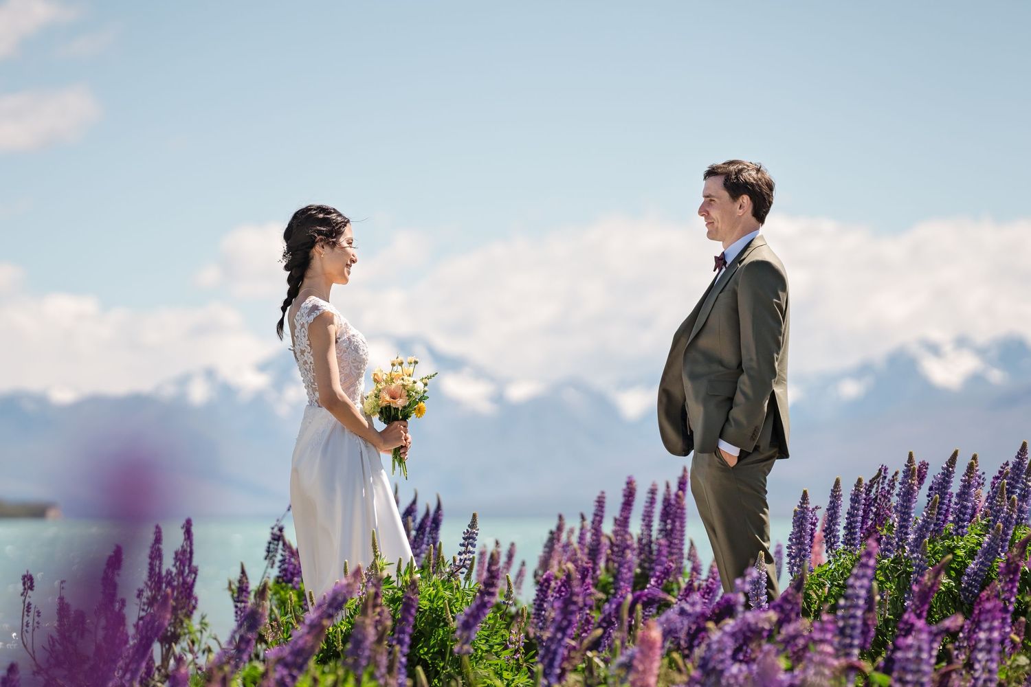 A couple stands in a scenic mountain meadow filled with vibrant purple lupine flowers on their wedding day.