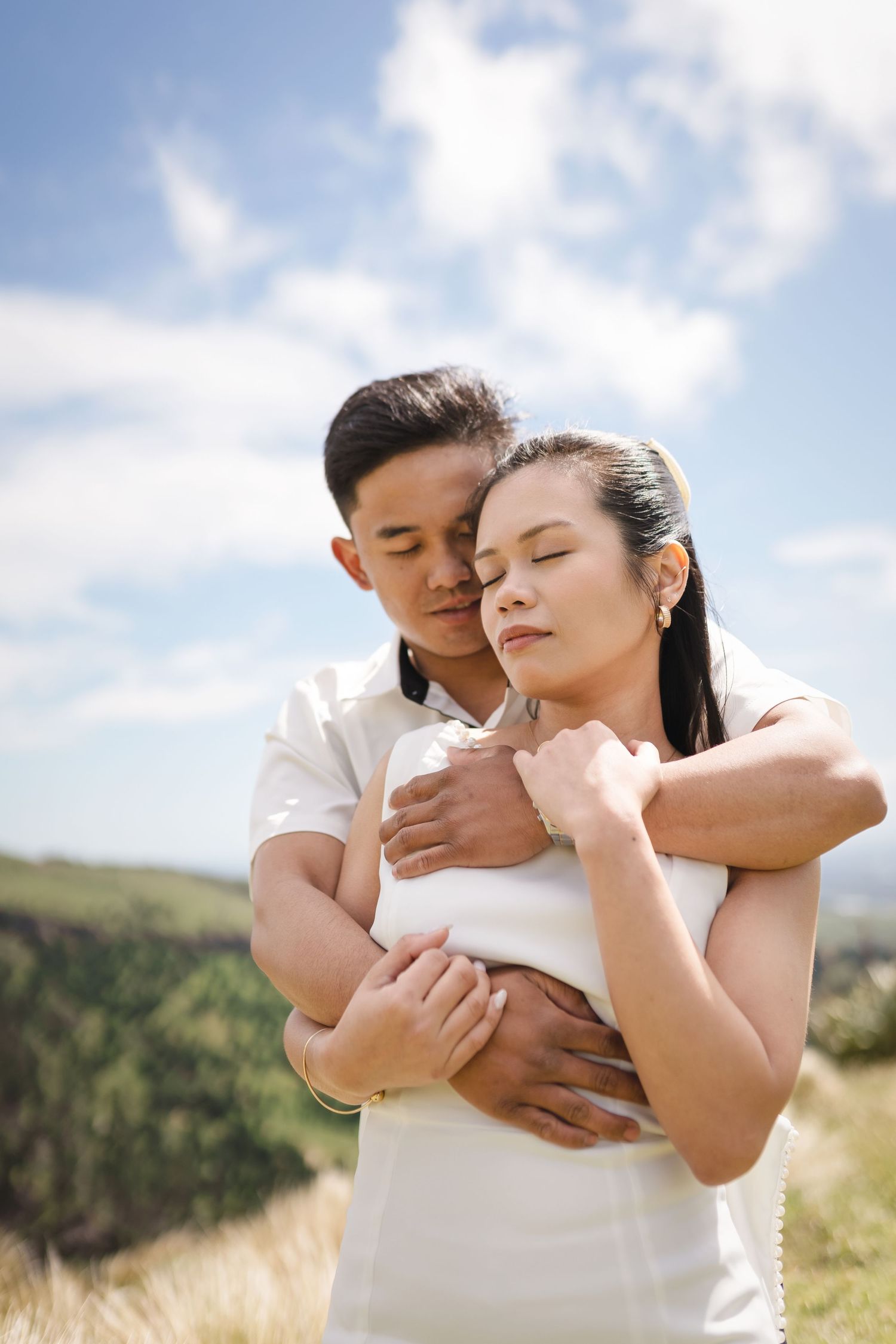 Couple embracing outdoors on a grassy hill, man hugging woman from behind with eyes closed under a blue sky.