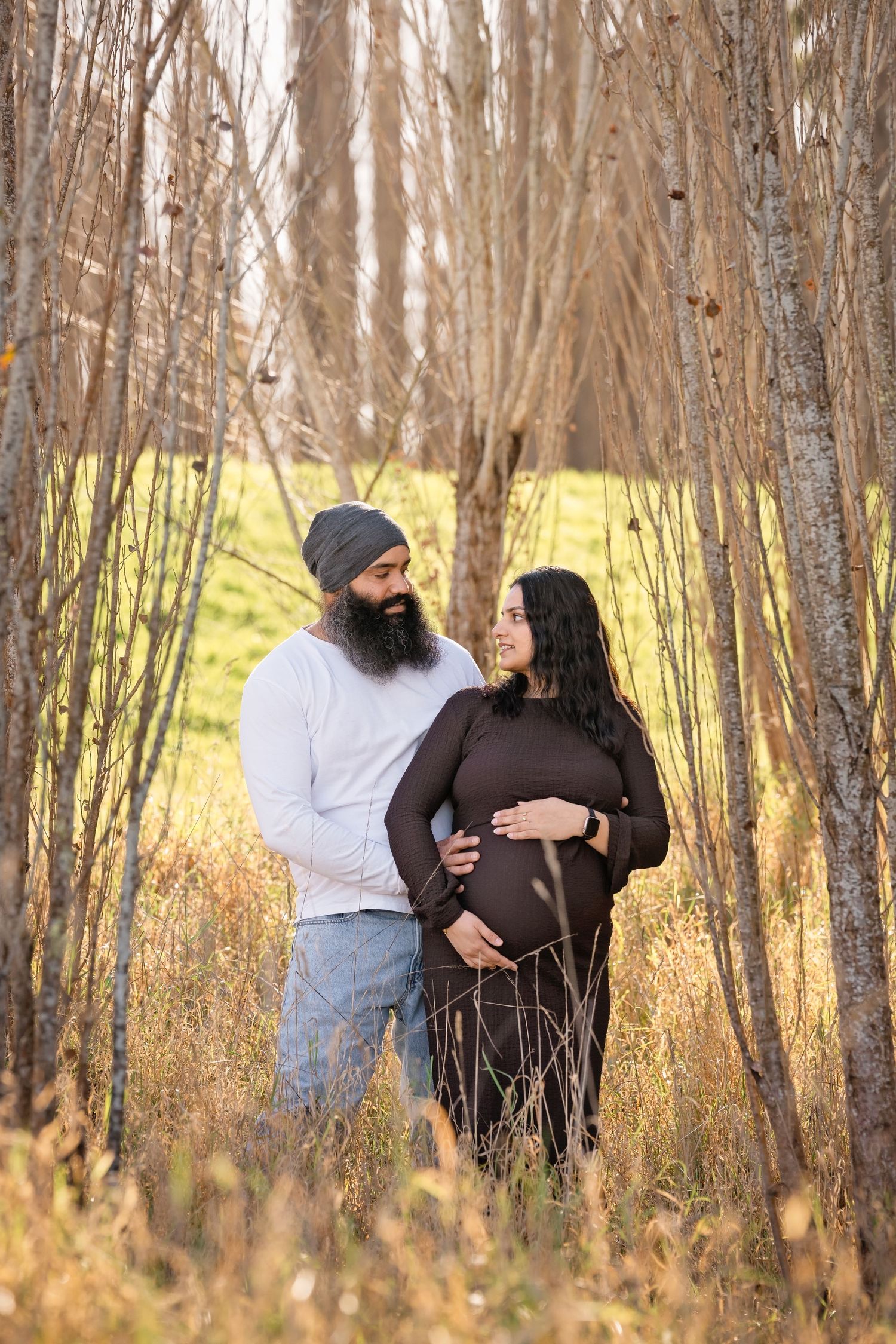 Expecting couple shares a tender moment among bare trees in a golden autumn forest setting.
