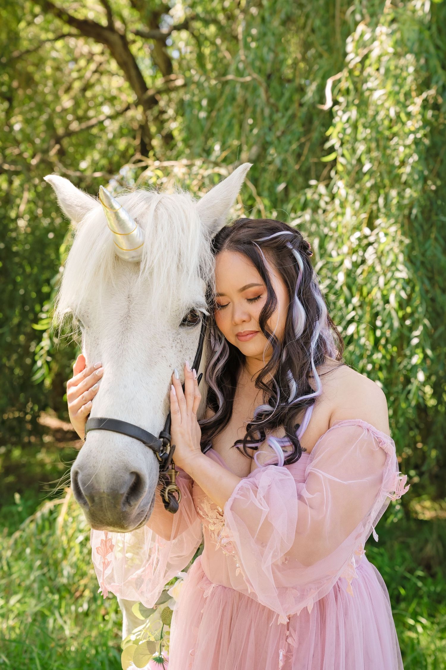 Young woman in pink dress tenderly embraces white horse with unicorn horn outdoors.