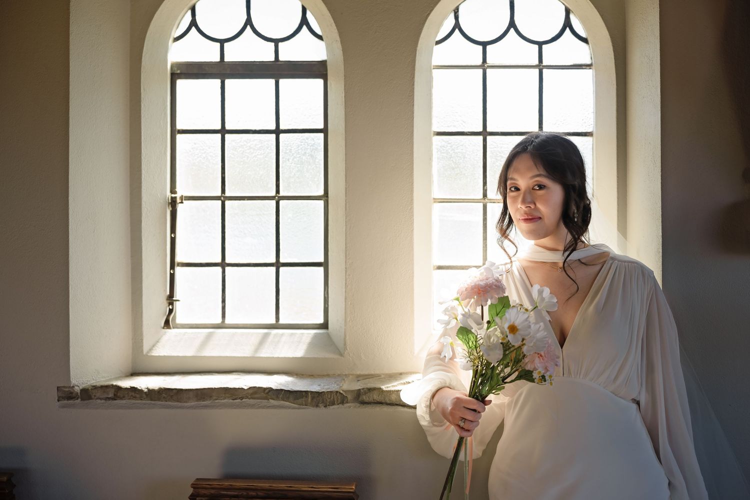 Young Asian woman in white dress holding flowers by arched windows in a sunlit church.