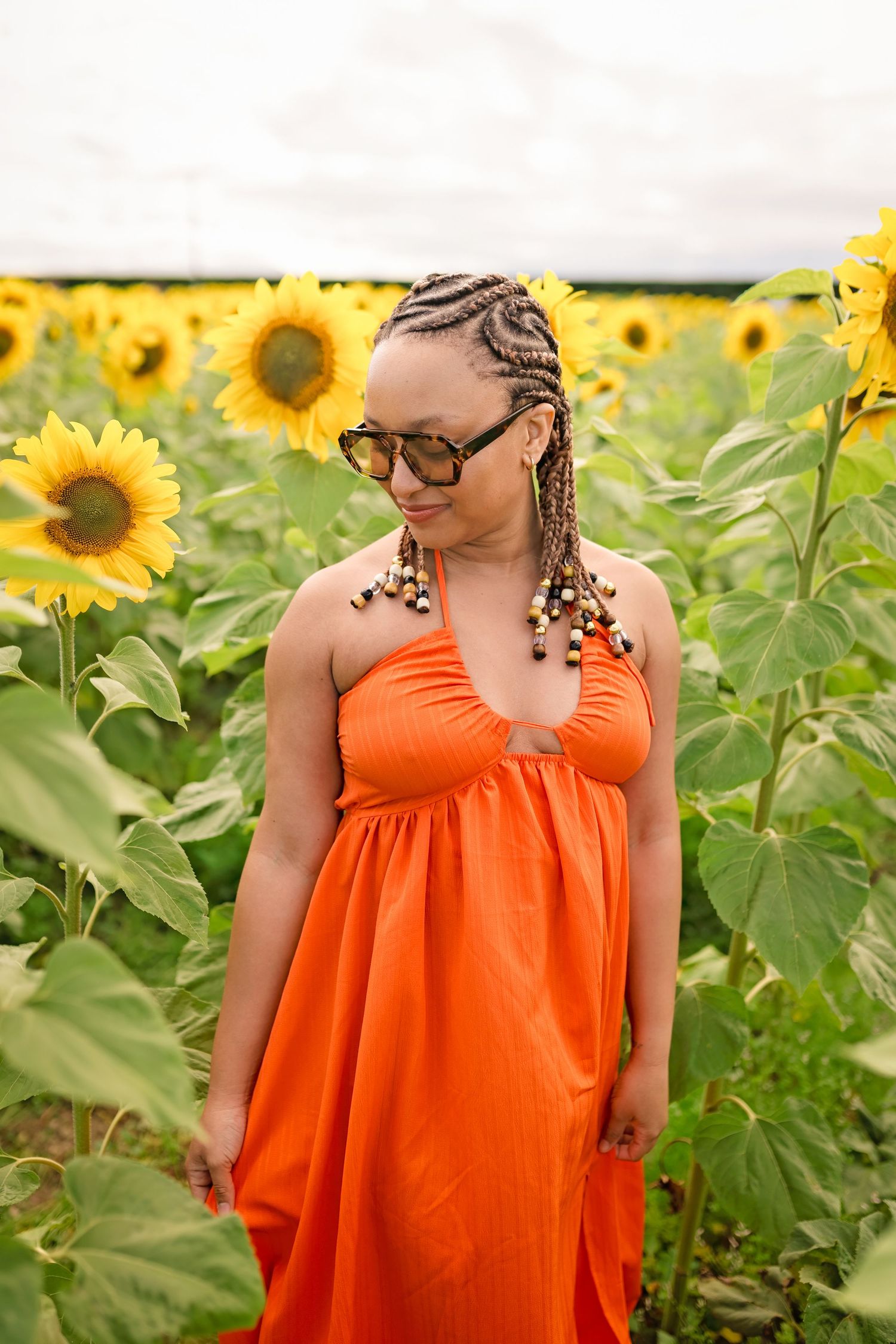 Woman in orange dress and sunglasses standing in sunflower field with braided hair adorned with beads.