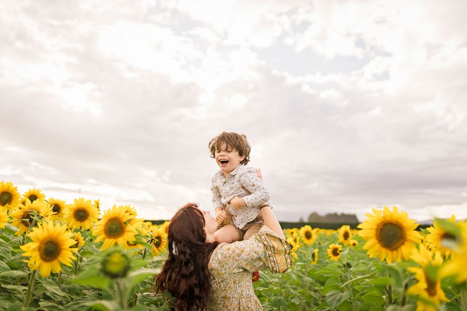A joyful mother lifts her laughing toddler above a vibrant sunflower field under a cloudy sky.