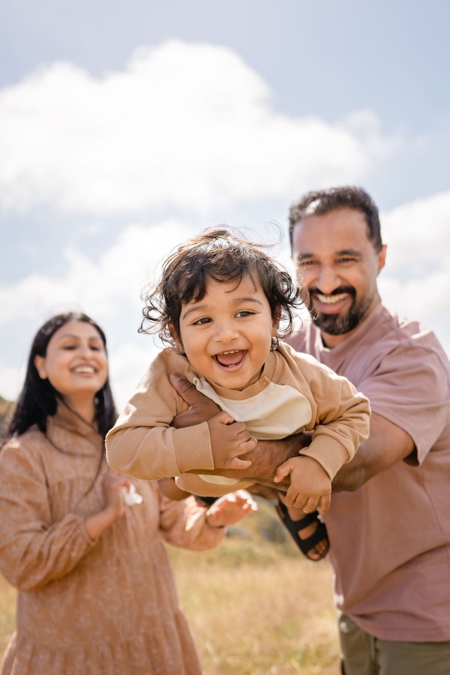 A joyful family outdoors, father lifting laughing toddler while mother smiles, set against a bright blue sky.