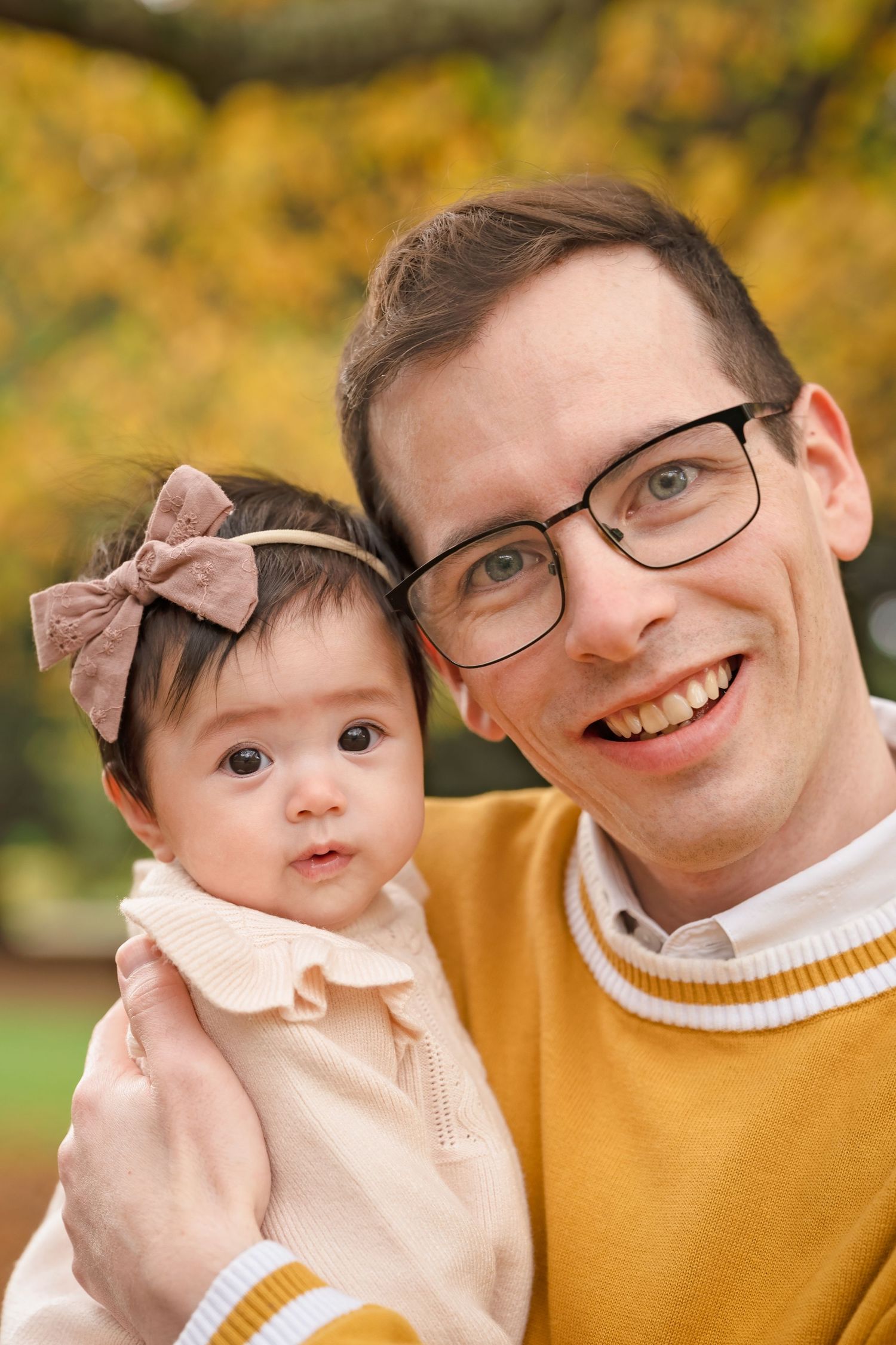Smiling man with glasses holding a baby girl with a bow headband in an autumn park setting.
