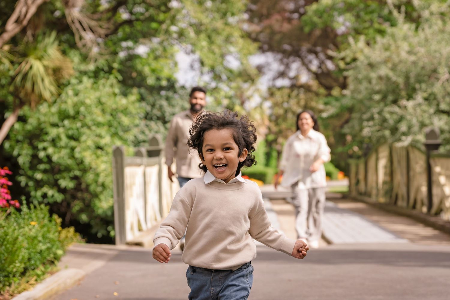 A joyful young child runs toward the camera in a sunny park while parents walk together in the background.