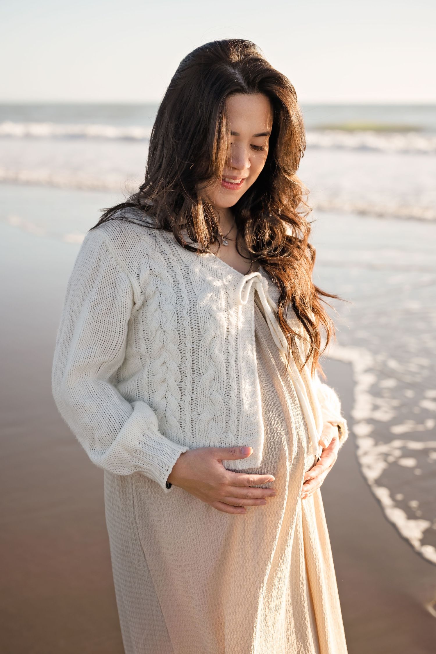 Pregnant woman in cream knit cardigan cradling her baby bump on a sunlit beach with ocean waves behind her.