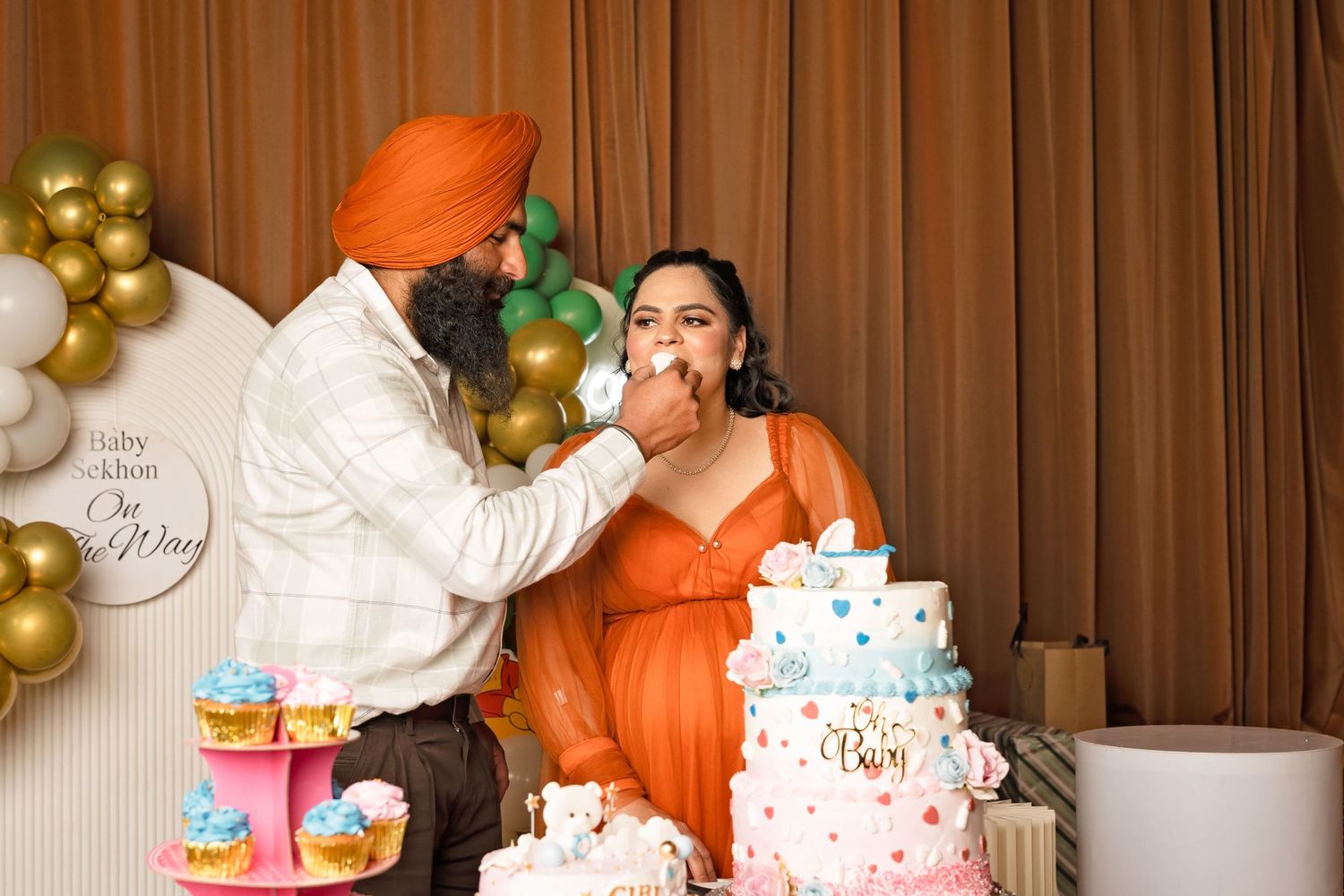 Sikh man feeding cake to pregnant woman at a baby shower with elegant tiered cake and balloon backdrop.