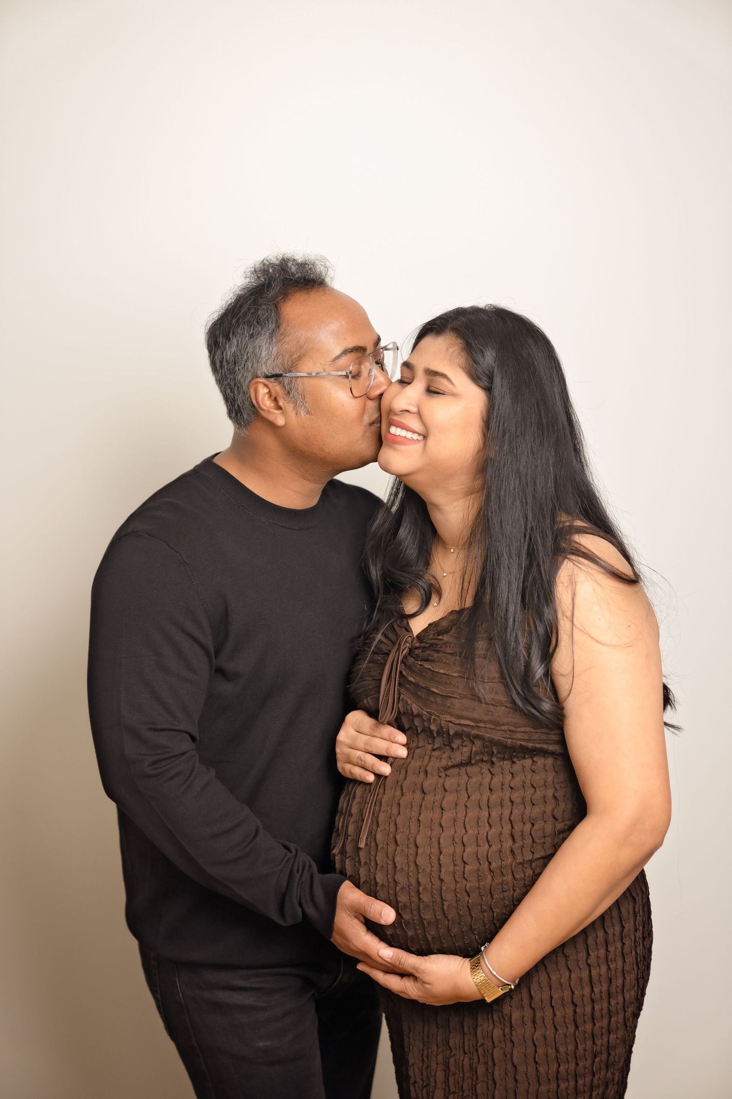 A couple in dark clothing share an intimate embrace and tender moment against a neutral background.