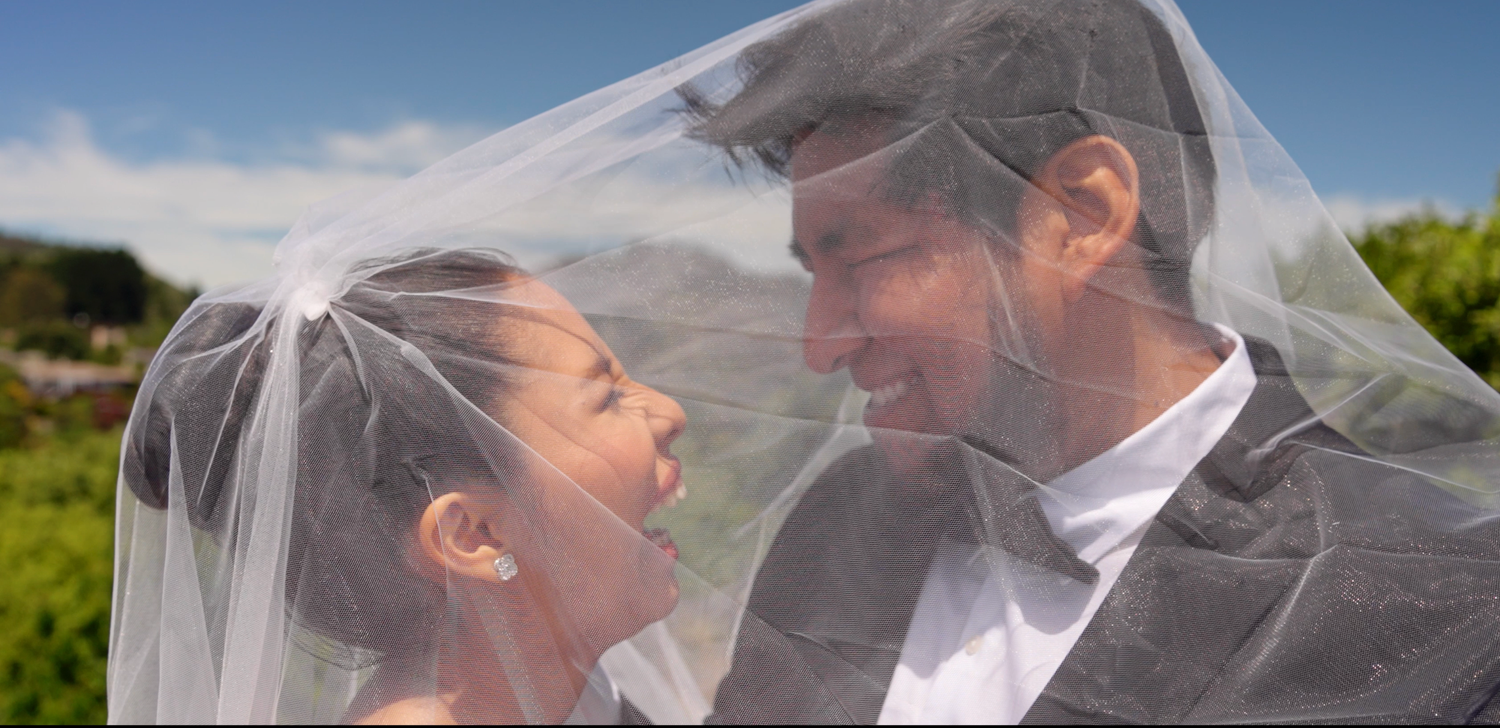 A happy couple shares an intimate moment on their wedding day under a white veil against a blue sky backdrop.