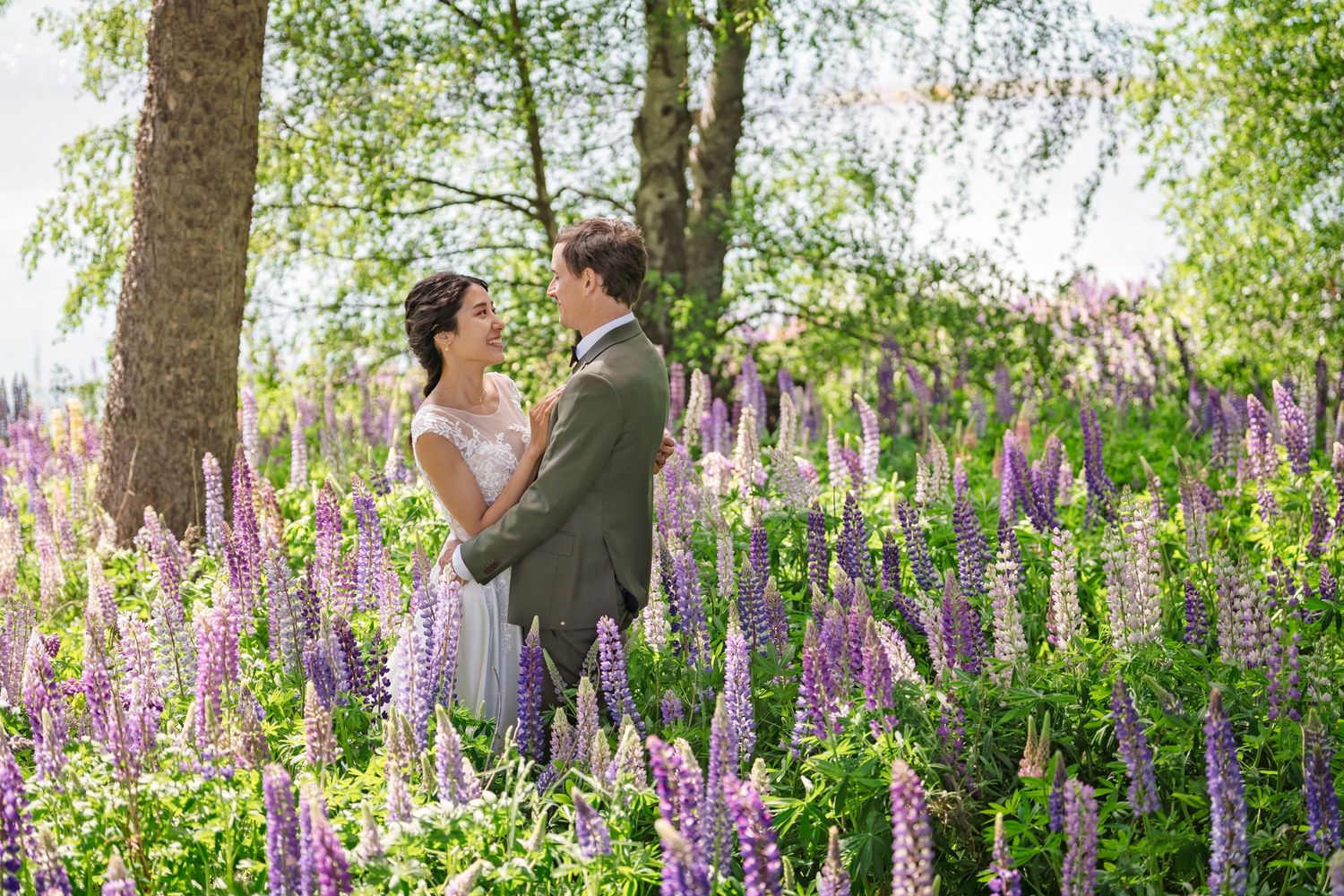 A couple stands together in a vibrant field of purple lupine flowers under a canopy of birch trees.