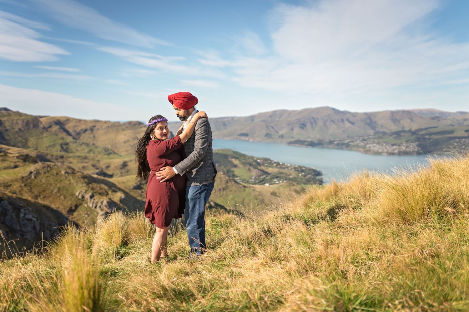 People stand on a scenic ridgeline hiking trail overlooking a tranquil bay and rolling mountains beneath blue skies.