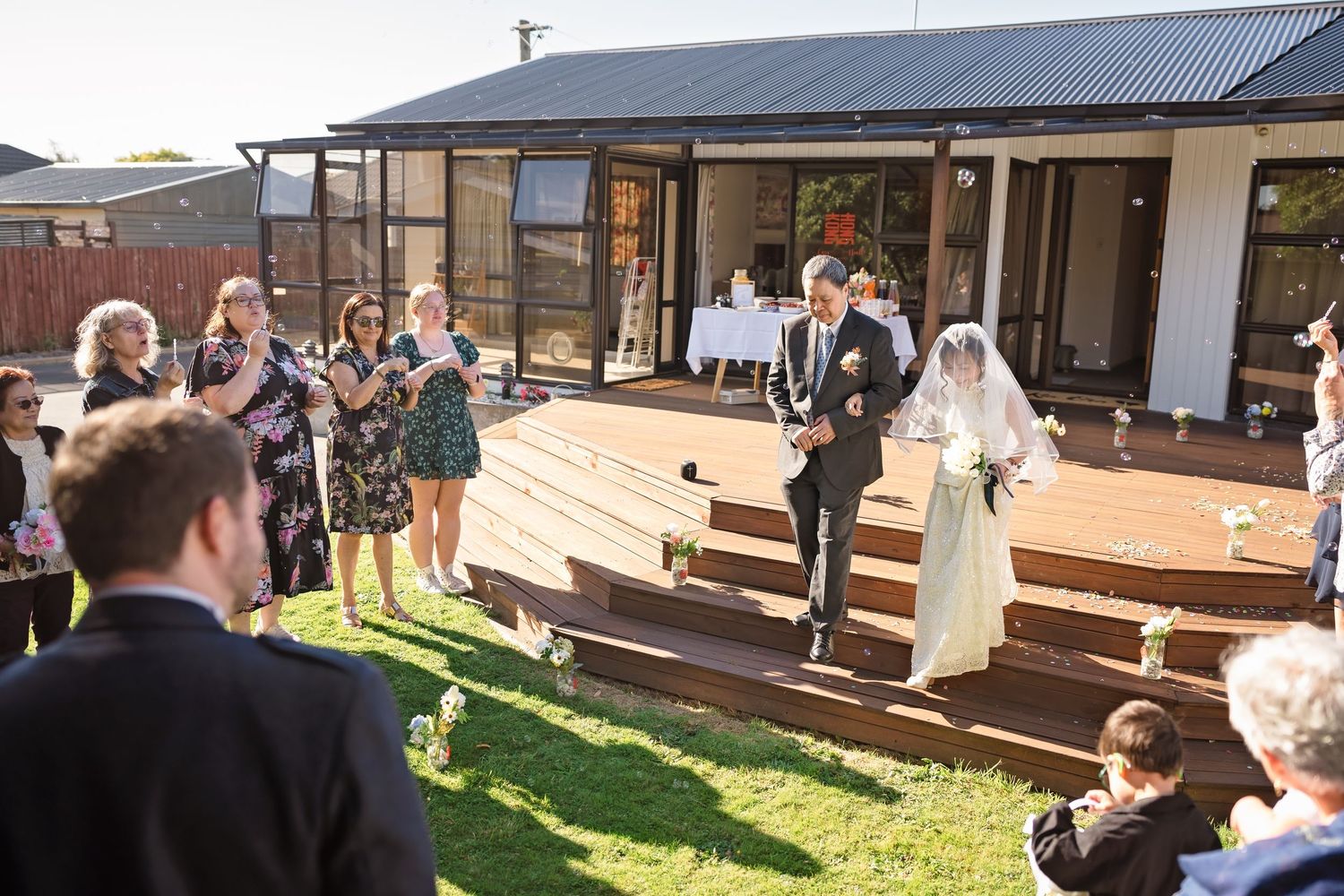 Bride and groom walk down outdoor aisle at backyard wedding ceremony with guests applauding on sunny day.