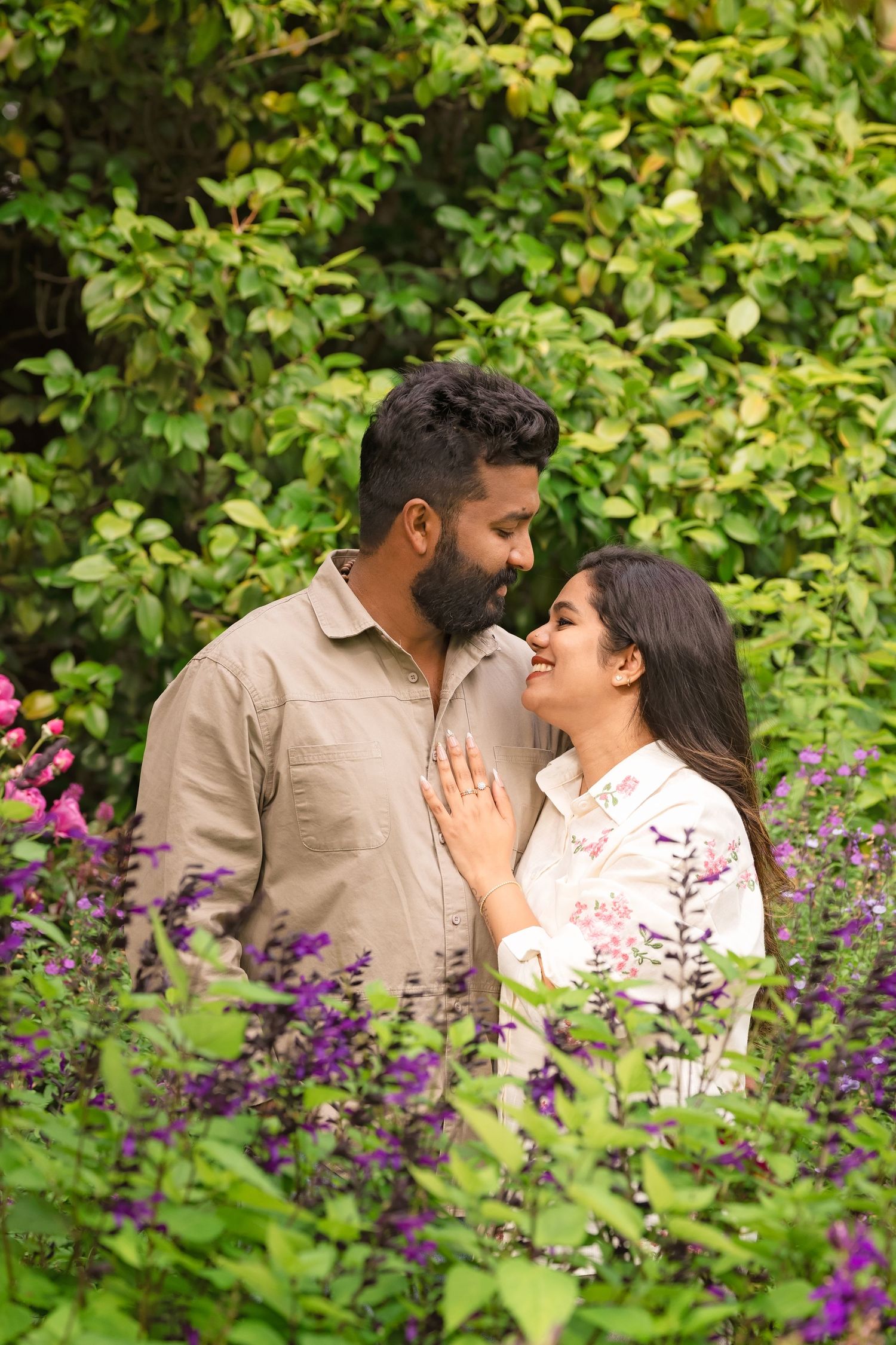 A couple shares a romantic moment surrounded by colorful flowers and lush green foliage in a garden setting.