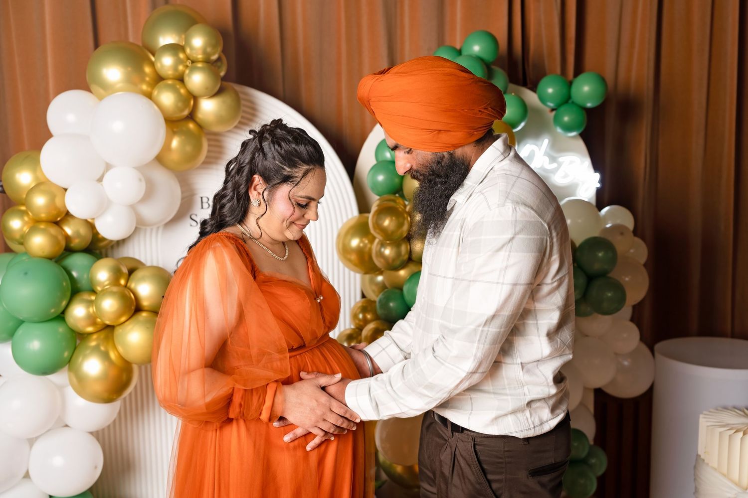 Sikh man in orange turban tenderly holding pregnant womans belly at decorated baby shower with colorful balloons.