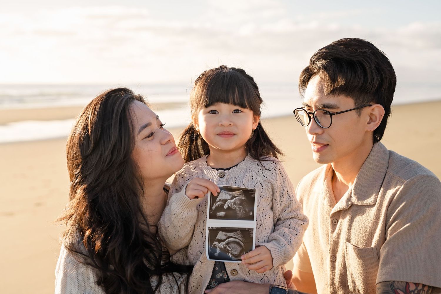 Family on beach, parents kissing toddler holding ultrasound photo, golden sunset backdrop.