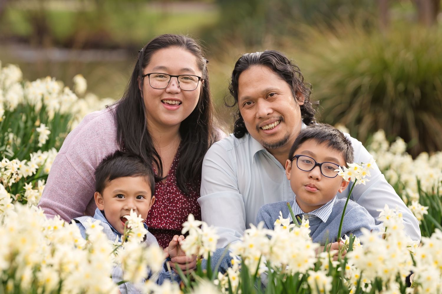 Family of four smiling among white daffodils in a garden, parents and two boys posing for a spring portrait.