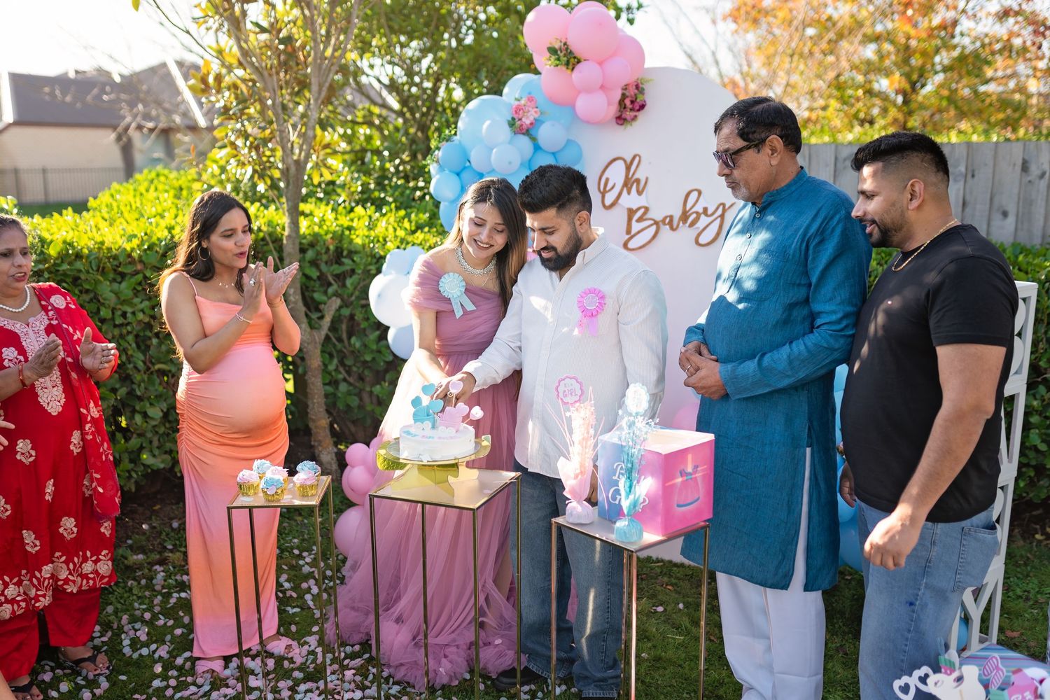 A gender reveal party celebration with pink and blue balloons being popped in a backyard setting during a sunny day.