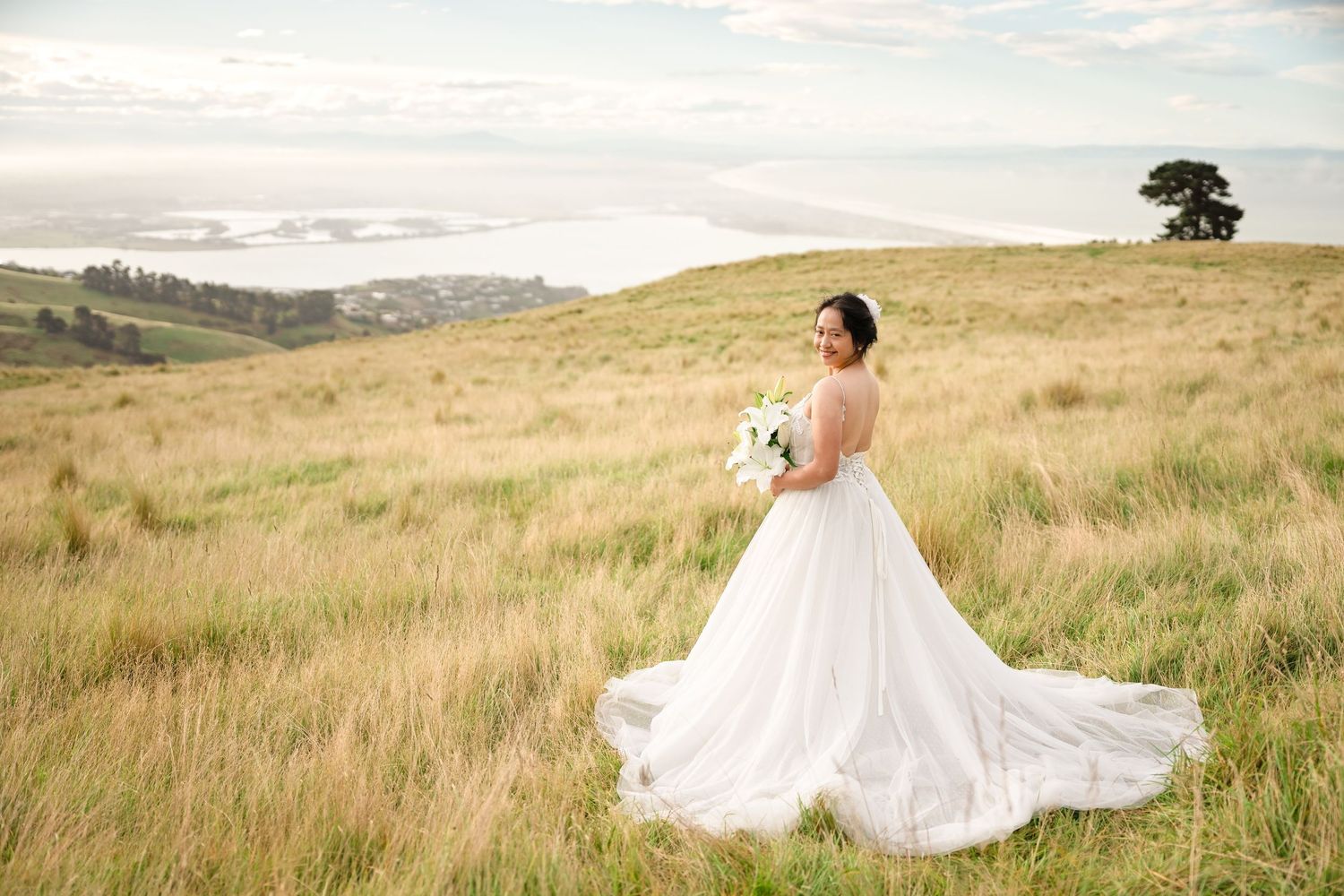 Bride in white ball gown holding bouquet stands in golden field with scenic coastal views behind her.