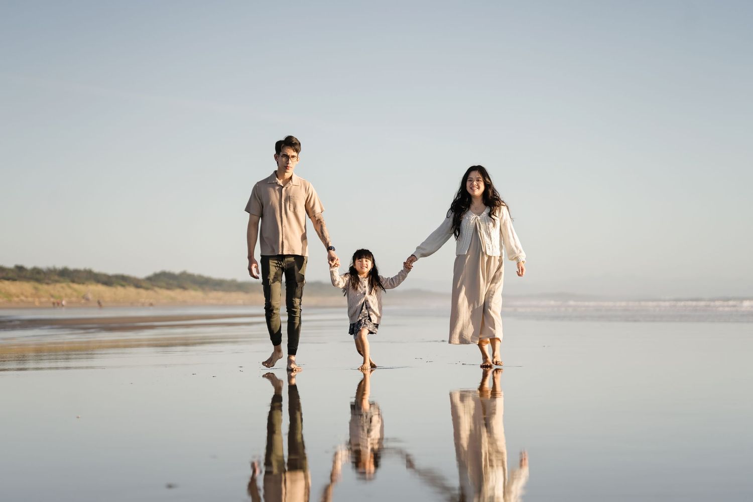 Family of three walking hand-in-hand on a reflective beach at sunset, parents swinging child between them.