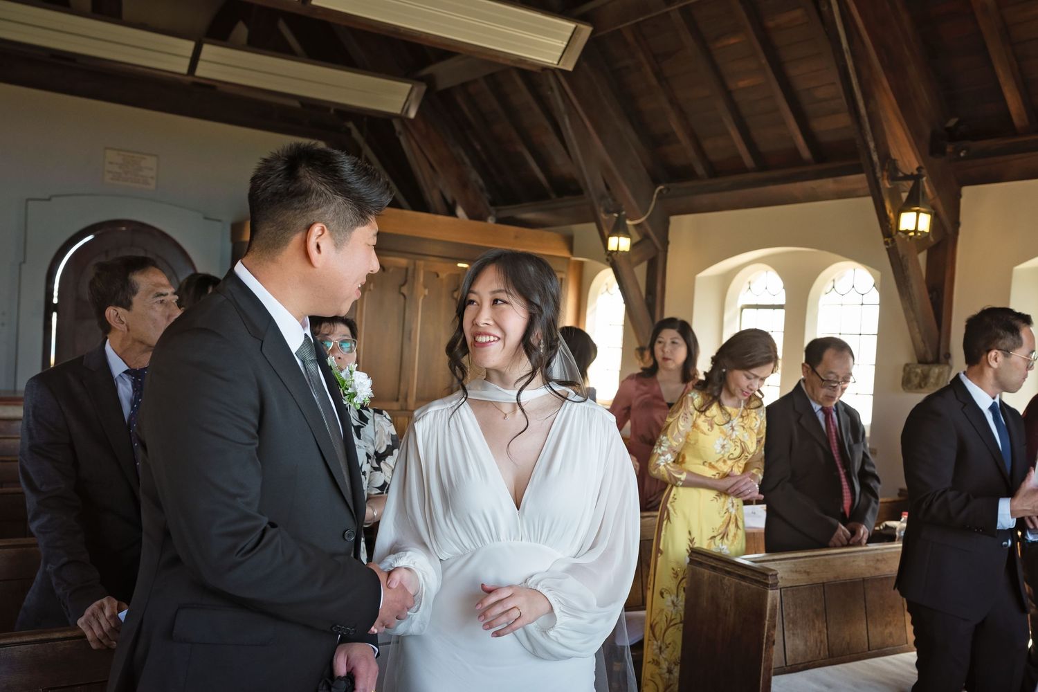 Bride in white gown smiles at groom inside rustic wooden chapel during intimate wedding ceremony.