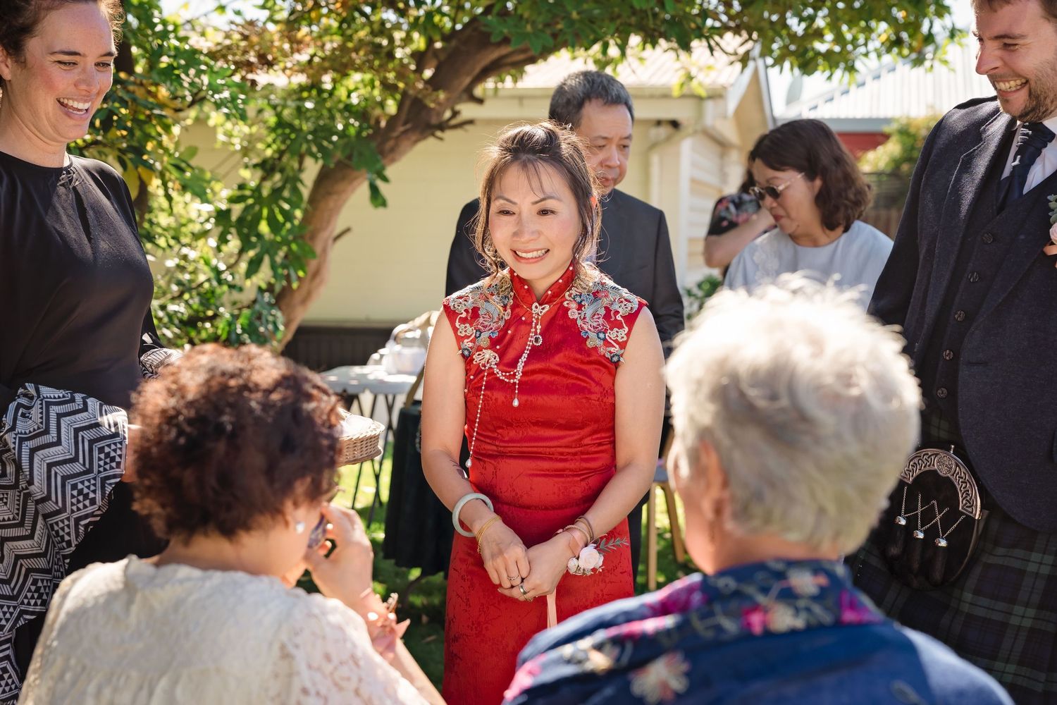 Woman in traditional red Chinese qipao dress smiles warmly while greeting wedding guests outdoors.