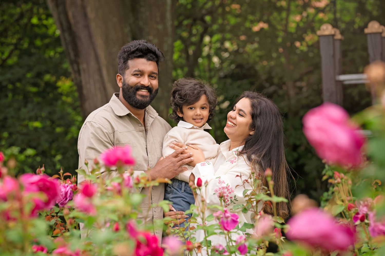 Family of three smiling among pink roses in a lush garden, parents holding toddler outdoors.